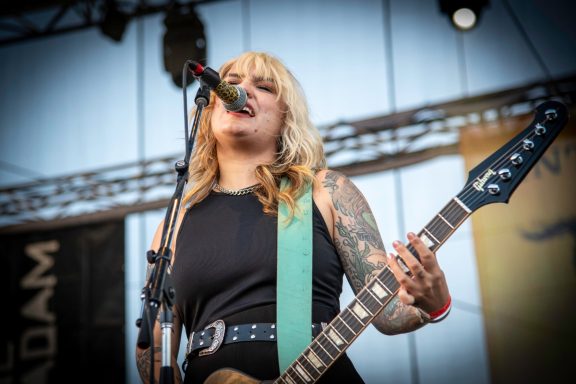 Female guitarist performing on stage, singing into a microphone, with tattoos visible on her arms.