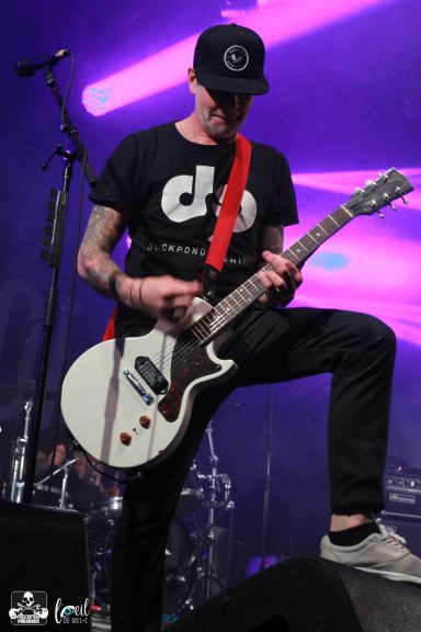 A musician playing electric guitar on stage, wearing a black t-shirt and cap.