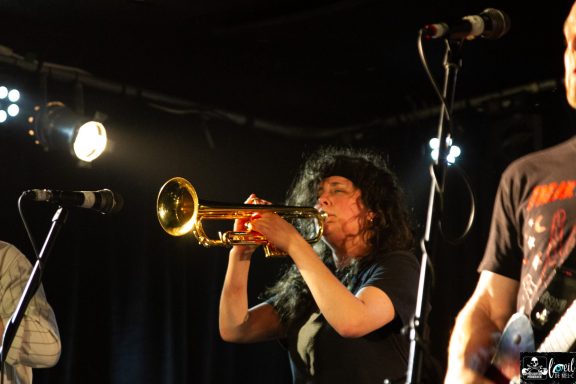Dead Bob - La Source de la Martinière Musician playing the trumpet on stage with a band, under bright stage lights.