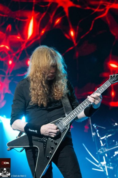 Musician with long curly hair playing guitar on stage with red and blue lighting.