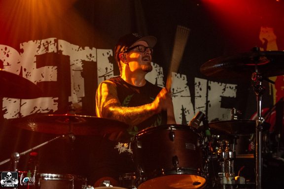 Drummer performing on stage with vibrant lighting and a band logo in the background.