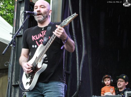 Musician performing with an electric guitar on stage, child and audience visible nearby.