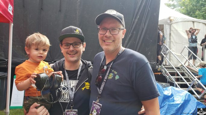 Two men pose for a photo, one holding a child, at an outdoor event.
