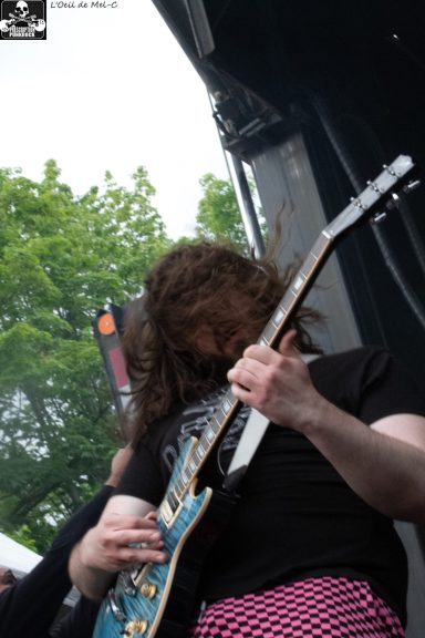 A guitarist performing passionately on stage, surrounded by greenery in the background.