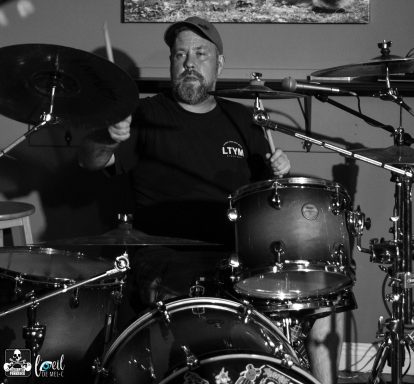 Drummer performing passionately on stage, surrounded by musical equipment.