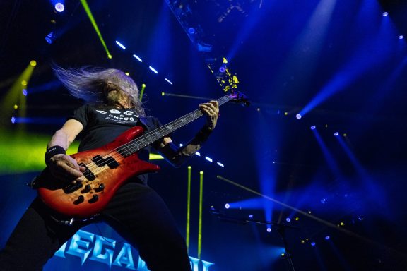 A musician performs energetically on stage with a red bass guitar and vibrant lights.