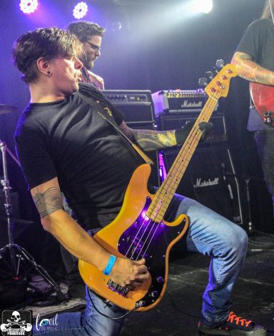 A musician performs on stage, playing an electric bass guitar with intense energy.