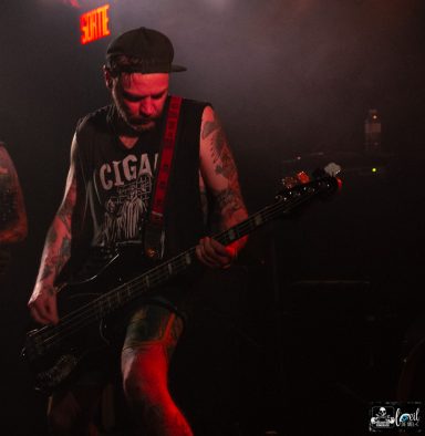 Straightline - L'Anti-Bar & Spectacles Musician playing bass guitar on stage, dressed in a sleeveless shirt and cap.