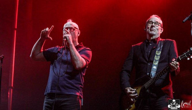 Two musicians performing on stage with dramatic red lighting. One is singing, and the other is playing guitar.