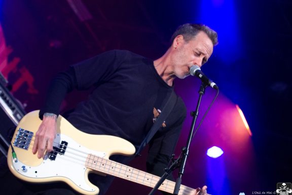 Musician playing bass guitar and singing on stage with colorful lighting.