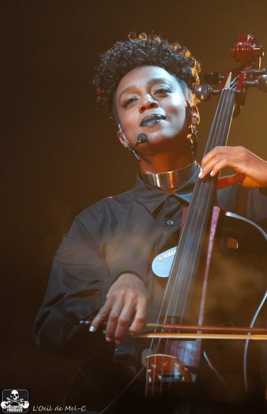 A musician playing the cello on stage, smiling with dramatic lighting behind.