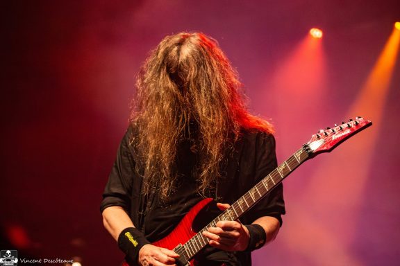 Musician playing electric guitar with long hair, performing on stage under bright lights.