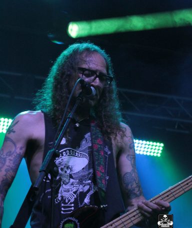 Musician with long hair and glasses performing on stage with a bass guitar.