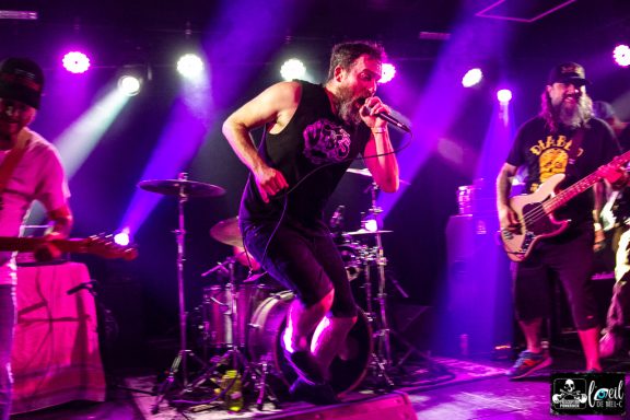 A band performing energetically on stage with vibrant purple lighting.