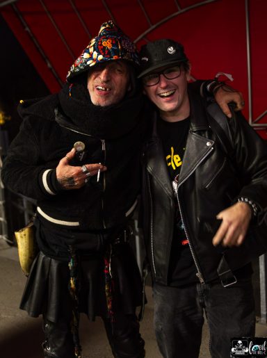 Two men smiling and posing together, one in a colorful hat and the other in a leather jacket.