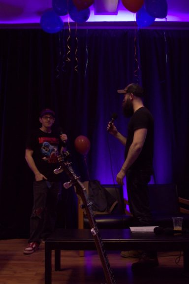 Two people stand in a dimly lit room with purple lighting and balloons, one holding a phone.