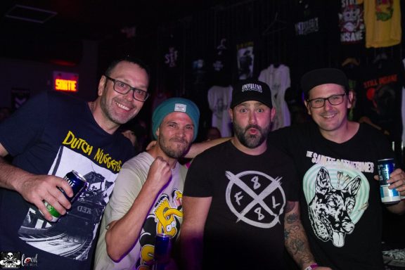 Four men standing close together, smiling, at a live music venue with merchandise in the background.