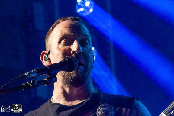 Musician performing on stage, singing into a microphone with blue lights in the background.