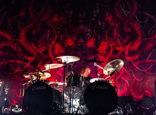 A drummer performing on stage with a vibrant red background featuring intricate designs.