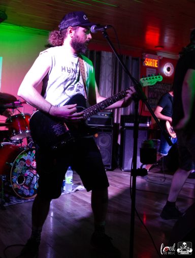 Man playing electric guitar on stage, wearing a tank top and shorts, with colorful lights in the background.