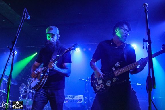 Two musicians perform on stage with guitars, illuminated by vibrant blue and green lights.