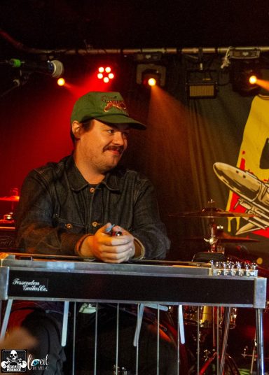 Musician playing steel guitar on stage, wearing a green cap and black jacket.