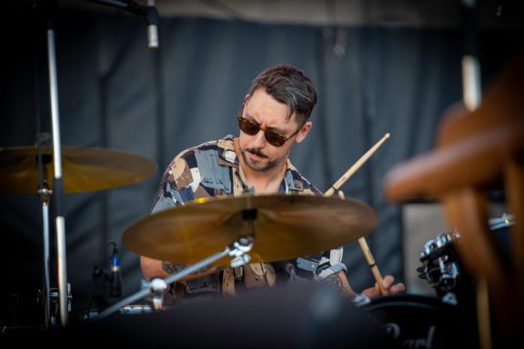Drummer in sunglasses playing a drum kit at a music event.