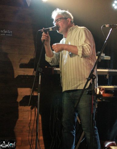 Dead Bob - La Source de la Martinière A musician performing on stage with a microphone, wearing a white shirt and glasses.