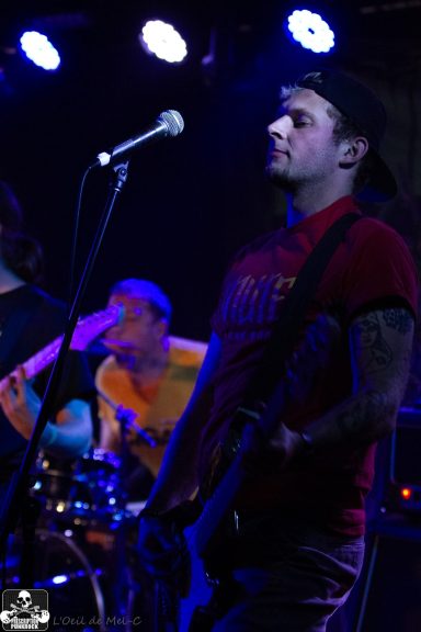 Band performing live on stage, with a guitarist in the foreground and musicians in the background.