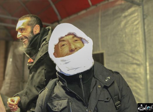 Two people indoors; one is smiling with a white scarf wrapped around their head.