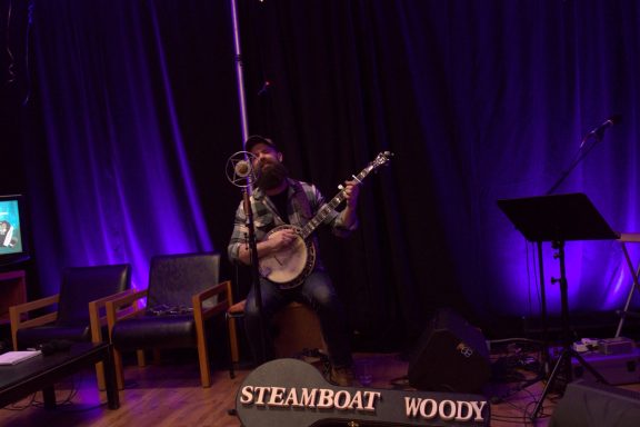 Musician playing banjo on stage with purple lighting and empty chairs in the background.