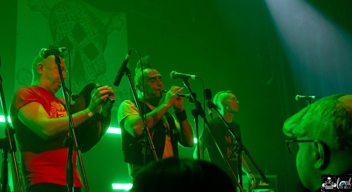 Three musicians perform on stage with green lighting and instruments.