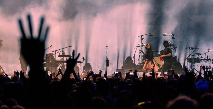 Crowd with raised hands at a concert, stage illuminated by dramatic lighting.