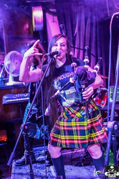 A musician in a plaid skirt plays the bagpipes on stage with a microphone.