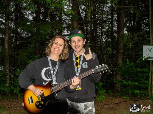 Two musicians pose outdoors, one holding a guitar and making a hand gesture.