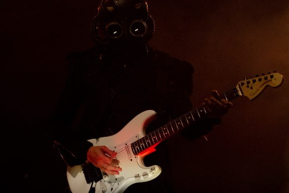 Person in a dark outfit playing an electric guitar with glowing eyes.