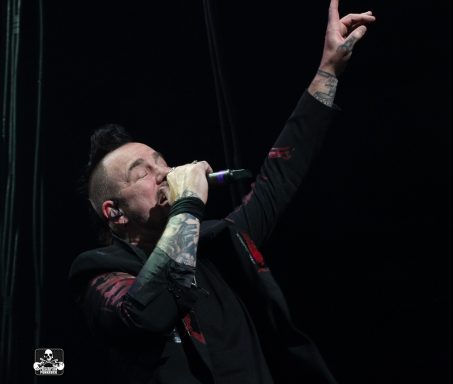A performer singing passionately with one hand raised and tattoos visible on their arm.