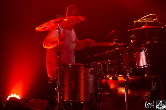 Drummer performing on stage with bright red lighting.