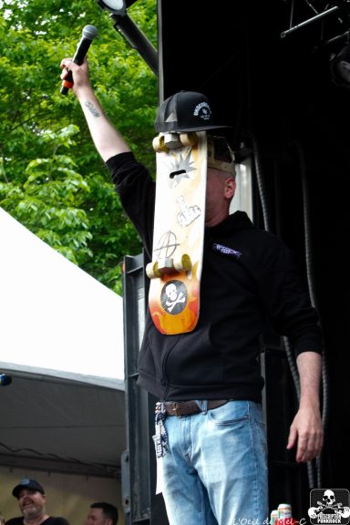 Person holding a skateboard overhead while wearing a hat, on stage at an outdoor event.