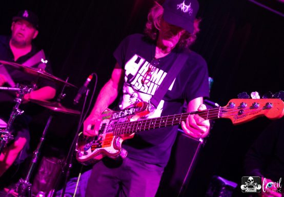 Musician playing bass guitar on stage with drummer in the background.