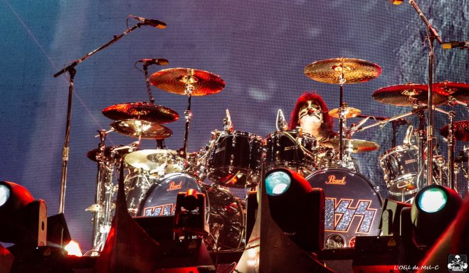 Drummer performing on stage with drums and cymbals, vibrant lighting in the background.