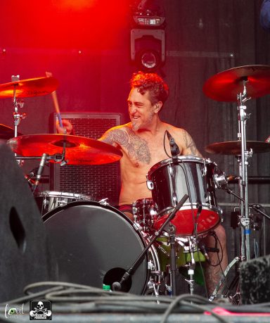 Shirtless drummer playing energetically on stage with vibrant lights behind him.