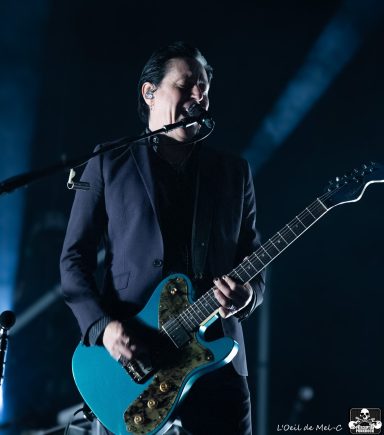 Musician performing on stage, holding a turquoise guitar, wearing a dark suit.