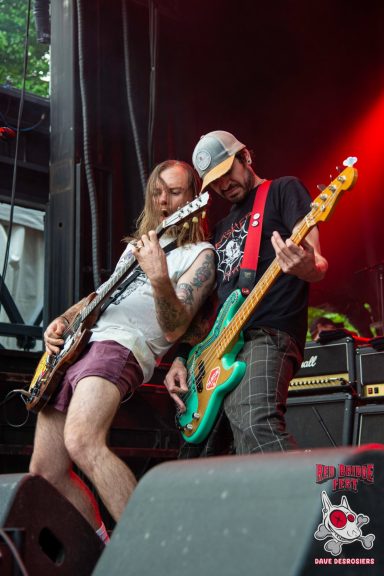 Two musicians perform on stage, one playing a guitar and the other a bass, with vibrant lighting.