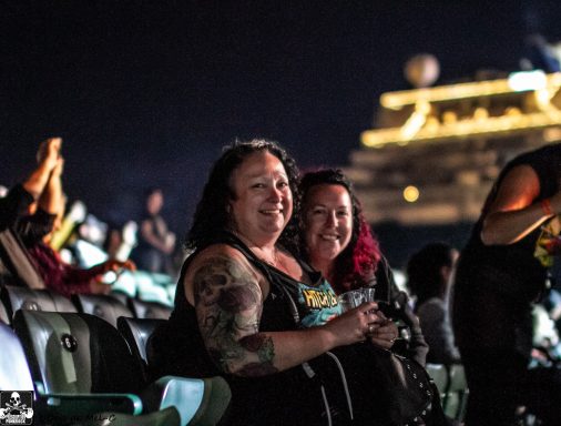 Two women smiling and sitting together in a crowded event space at night.