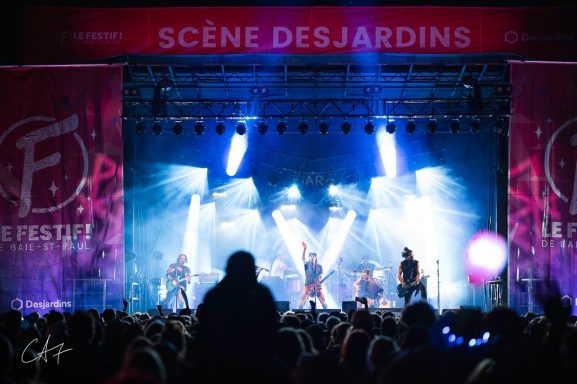 Band performing on stage with bright lights and a large audience.