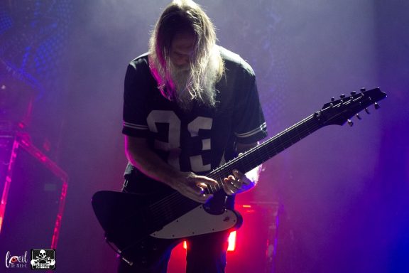 Musician with long hair playing a guitar under colorful stage lights.