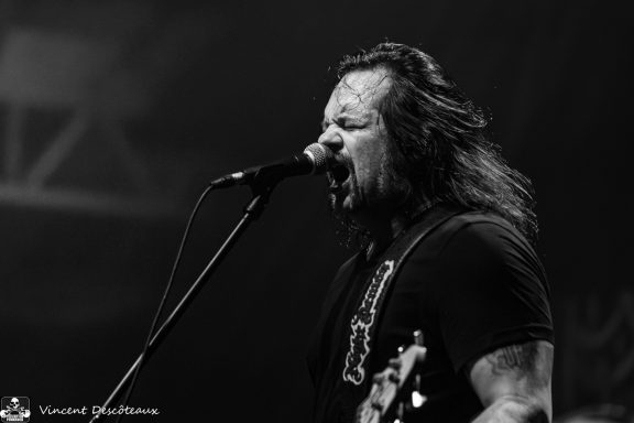 Singer with long hair performs passionately into a microphone on stage.