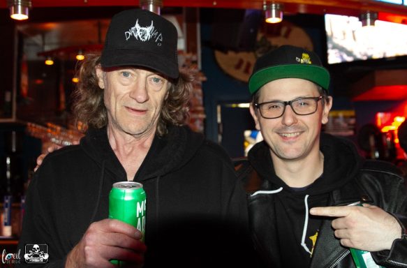 Two men posing together, one holding a green can, in a bar setting.