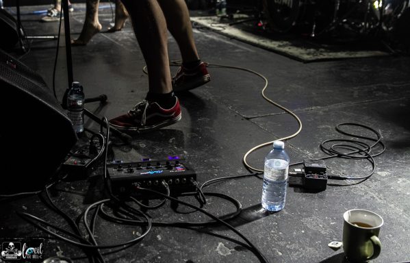 Straightline - L'Anti-Bar & Spectacles Feet in sneakers on a stage with cables, a water bottle, and a coffee cup nearby.
